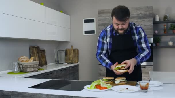 Homme barbu épais ou gras cuisine un hamburger fait maison dans une cuisine moderne — Video