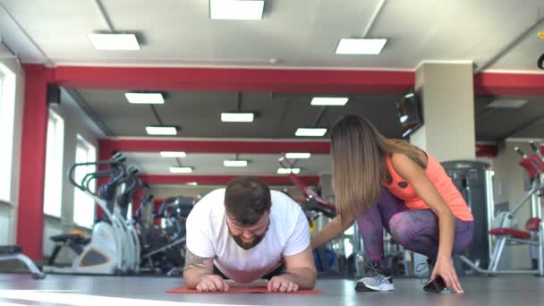 Ein kaukasischer Mann mit Bart führt eine Übung in der Turnhalle Plank, neben schönen Mädchentrainer, Training — Stockvideo