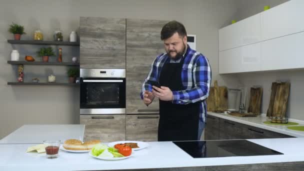 Kalın sakallı erkek gıda ev yapımı hamburger pişirmeden önce modern bir mutfak, bir telefonu kullanarak bir fotoğrafı çeker — Stok video