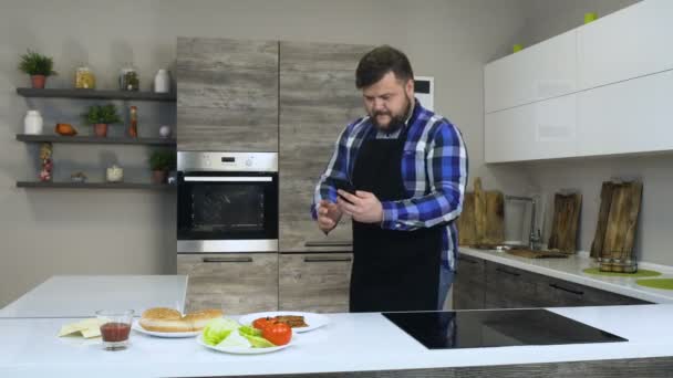 Dikke bebaarde man neemt een foto van voedsel met behulp van een telefoon in een moderne keuken, vóór het koken van een zelfgemaakte Hamburger — Stockvideo