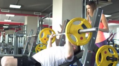Sakallı şişman beyaz adam bir halter bench press egzersiz sırt üstü bir güzel kız eğitmen, göğüs kasları güvenliğini sağlama sırasında gerçekleştirir