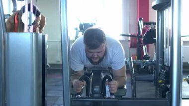 Sakal ile bir şişman beyaz adam bir bacak kas egzersiz bireysel bir eğitmen ile spor salonunda meşgul, dizlerinin içinde bacaklar bükme, egzersiz