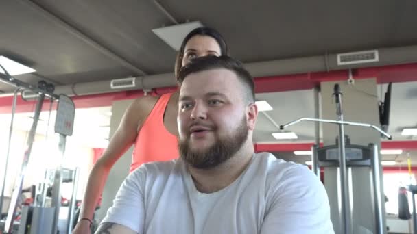 Erwachsener Mann mit Bart ist im Fitnessstudio beschäftigt, um mit einem Personal Trainer Mädchen die Brustmuskulatur am Kamerasimulator zu trainieren, Fitness — Stockvideo