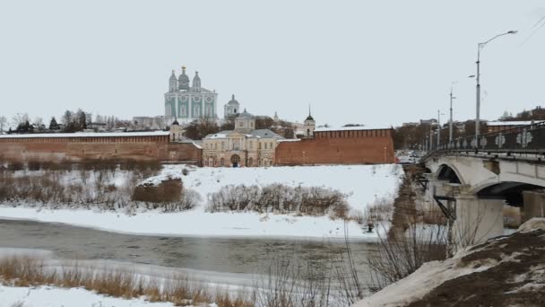 V zimě protéká řeka, na břehu se nachází palác, pravoslavný kostel, pevnost s červenými zdmi v russaském městě. Na ulici je sníh, Starý most s černými zábradáči a lampami — Stock video