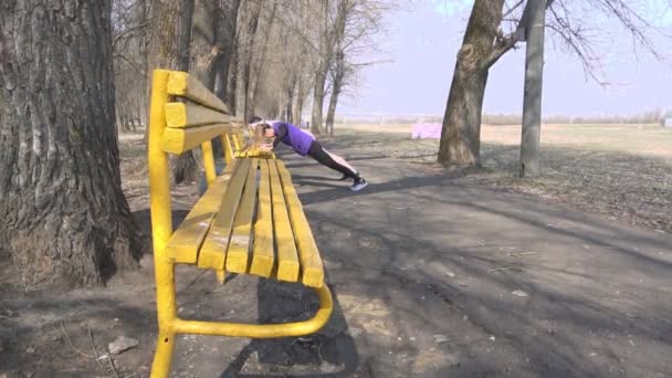 Homem atlético com barba e uma treinadora praticam esportes no parque sobre a natureza de fazer os exercícios flexões do banco, câmera lenta — Vídeo de Stock