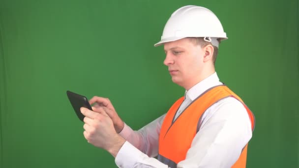 A male engineer in a protective helmet and signal vest stands with a tablet on a construction site, makes notes, modern technologies, green background, chromakey — Stock Video