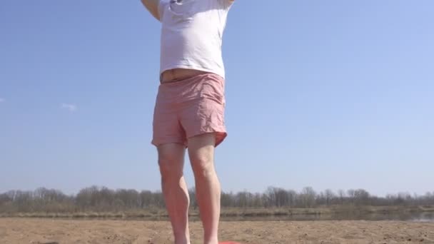 Dicker kaukasischer Mann in weißem T-Shirt mit Bart, der dynamisches Yoga in der Natur an einem Sandstrand betreibt, sich mit den Händen dreht, Selbstverbesserung — Stockvideo