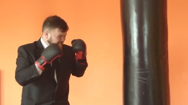 Joven hombre caucásico guapo con una barba en traje. Un empleado de oficina. El gerente maneja su ira en el gimnasio. Bolso de boxeo, cámara lenta, fondo — Vídeo de stock