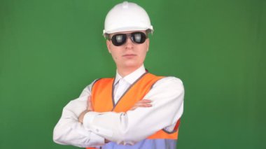 Siyah gözlüklü, koruyucu beyaz kasklı ve kameraya bakan sinyal gileti ile beyaz renkli genç bir adam, yeşil arka plan, hromakey, kazaların önlenmesi