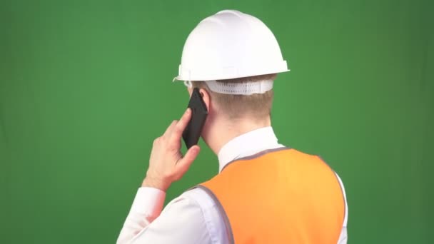 Un hombre con un casco protector blanco y chaleco de señal hablando por teléfono en un sitio de construcción, fondo verde, hromakey, industria, profesional — Vídeos de Stock