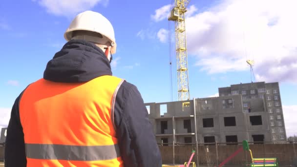 Bir kask ve sinyal yeleği siyah gözlüklü bir erkek mühendis yazıyor ve çok katlı bir konut binasının bir inşaat alanında bir tablet işaretleri, kopya alanı, yakın çekim — Stok video