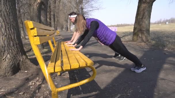 Друзья юная девушка и мужчина занимаются спортом весной на природе в парке, отжимания со скамейки, силовые тренировки, медленное мо — стоковое видео