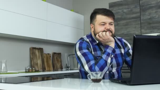 Товстий бородатий хлопець дивиться на ноутбук. Людина з надмірною вагою насолоджується кавою, сидячи на кухні вранці. Товстий чоловік дивиться стрічку новин в блокноті і серфінгах в Інтернеті, спілкуючись з послідовниками або — стокове відео
