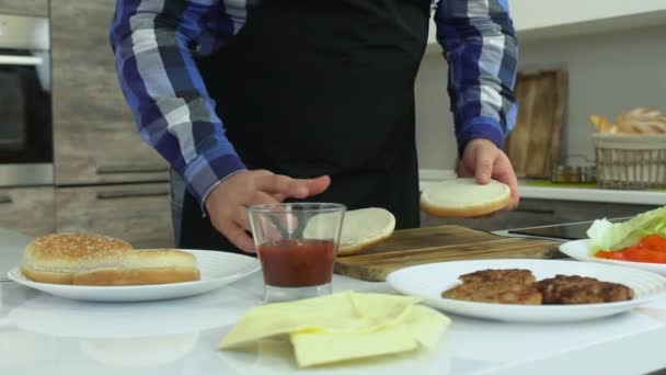 Hustý chlápek vaří hamburgery doma. Buclatý muž poskvrní rajčatovou pastu na housku. Zdravý životní styl, smažené a vysoce kalorické škodlivé potraviny. Riziko obezity a nadváhy. Zpomaleně. — Stock video