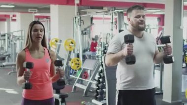 Bireysel antrenör ile kalın obez adam spor salonunda dumbbells ile fiziksel jerks yapmak. Kilolu adam ve kadın eğitmen kalori yakmak. Fitness egzersizi yapan kişisel eğitmen ile Tombul erkek
