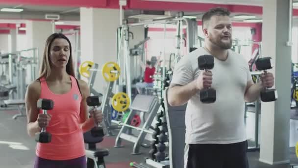 Thick obese guy with individual coach do physical jerks with dumbbells in gym. Overweight man and female trainer burn calories. Plump male with personal instructor doing fitness exercise — Stock Video
