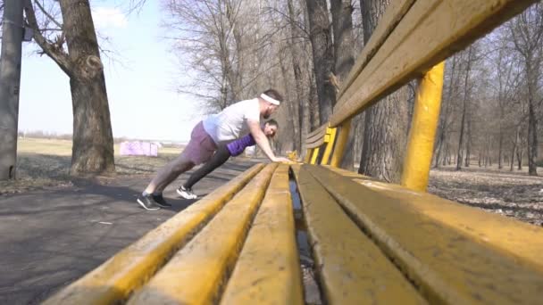 Sportowiec kaukaski mężczyzna z brodą i dziewczyna trener sportu grać w parku na charakter robi ćwiczenia push-up z ławce, zwolnionym tempie — Wideo stockowe