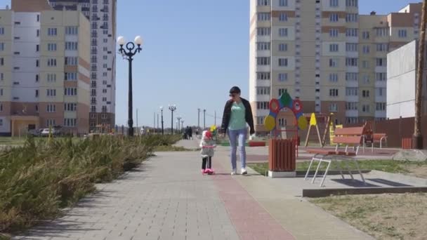 Hermosa chica caucásica madre enseña a su hija pequeña a montar un scooter en el parque en el callejón, la infancia — Vídeo de stock