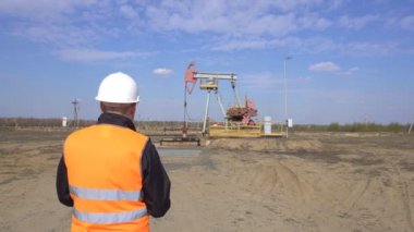 Sinyal yeleği ve beyaz kasklı bir erkek müfettiş-operatör, pompalama ünitesi petrol, gaz ve yanıcı yakıt, sanayi, fotokopi alanı, pumpjack hakkında okumaları kaydeder