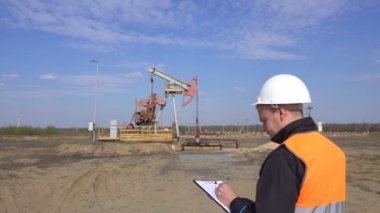 Sinyal yeleği ve beyaz kasklı bir erkek müfettiş-operatör, pompalama ünitesi petrol, gaz ve yanıcı yakıt, endüstri, fotokopi alanı, pumpjack, mühendislik hakkındaki okumaları kaydeder.