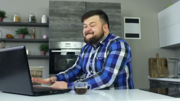 Um cara grosso está na cozinha na frente de um laptop e começa a ficar com raiva, bate a mesa com a mão. Um homem gordo senta-se na cozinha em frente a um laptop e começa a ficar com raiva, bate a mesa — Vídeo de Stock