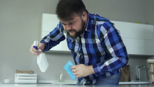 El hombre con sobrepeso rocía su detergente sobre la mesa. Chico hinchado limpia su cocina con servilleta de la suciedad, grasa y manchas en la superficie sucia. Gordo macho en jeans y camisa con paño esponjoso. Movimiento lento — Vídeo de stock