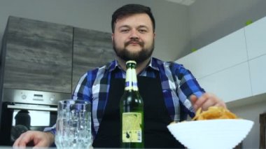 Şişman adam masada mutfakta oturur ve bir bıçakla bir şişe bira açar. Patates cipsi tabakta. Alkollü içecek ve sağlıksız yüksek kalorili zararlı gıda. Obezite riski ve