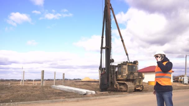 O engenheiro principal em copos pretos, um colete de sinal e um capacete falam no telefone contra o contexto do começo da construção de uma nova casa em pilhas, equipamento para dirigir em pilhas, um diesel — Vídeo de Stock