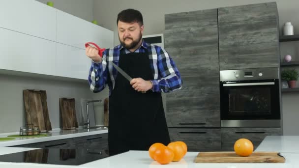 A fat bearded man in an apron stands in the kitchen and sharpens a knife in front of the table with oranges. A big male whets a knife — Stock Video
