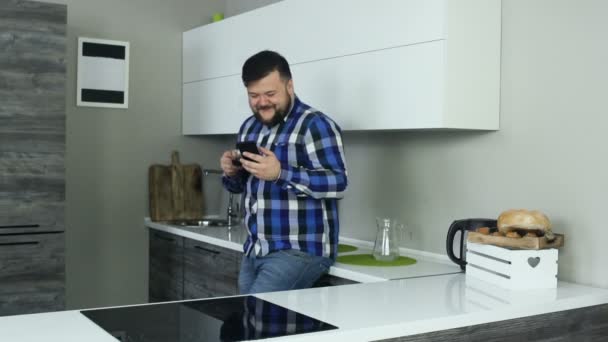 Ένας εύσωμος άντρας στέκεται στην κουζίνα και γελάει, κοιτάζοντας το τηλέφωνο, και μετά πίνει μια γουλιά καφέ. Ένας χοντρός είναι στην κουζίνα και γελά όταν κοιτάζει το smartphone, και μετά πίνει καφέ.. — Αρχείο Βίντεο