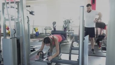 Özel kadın antrenör spor salonunda simülatör üzerinde egzersiz yapmak nasıl adam gösterir. Adam bireysel eğitmenle bacak kaslarını sallayacak. Fitness kulübünde kişisel antrenör kilo kaybı matkaplar