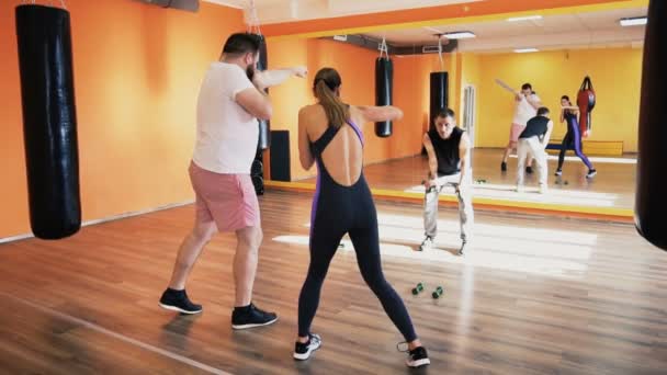 Personal training in fitness club. Private coach explains to a fat man blows with dumbbells. A slim girl coaches sharp kicks with strenght exercises. Individual weight loss drills for thick guy. Warm — Stock Video