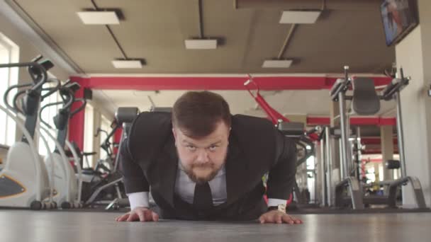 Un homme d'affaires caucasien masculin en costume noir effectue des exercices physiques dans une salle de gym, se tordant hors du sol et regarde dans la caméra comme un concept d'un mode de vie sain employés de bureau — Video