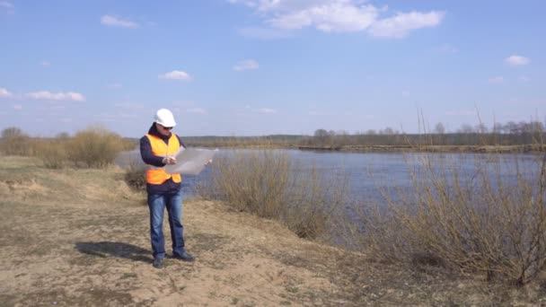 Mannelijke ingenieur in een signaal vest en een helm kijkt naar een ondiep waterplan tegen de achtergrond van een rivier, industrie, inspecteur — Stockvideo