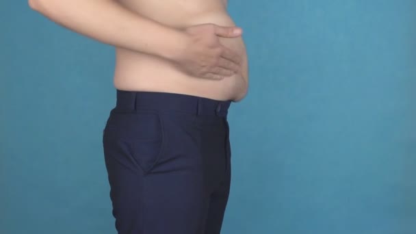 Ένας νεαρός άντρας κλίνει πάνω από μια παχιά κοιλιά λίπους με υπέρβαρος σε μπλε φόντο, η έννοια των προβλημάτων με την παχυσαρκία και την κυτταρίτιδα, αργή mo — Αρχείο Βίντεο