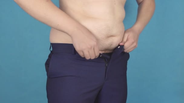 Gordo joven con un gran vientre se abrocha los pantalones apretados en su cintura concepto de sobrepeso y obesidad, fondo azul, abdomen — Vídeo de stock