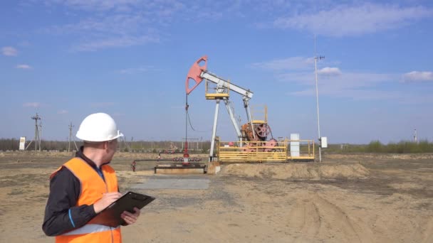 Sinyal yeleği ve beyaz kasklı bir erkek müfettiş-operatör, pompalama ünitesi petrol, gaz ve yanıcı yakıt, sanayi, fotokopi alanı, pumpjack, petrol ile ilgili okumaları kaydeder. — Stok video