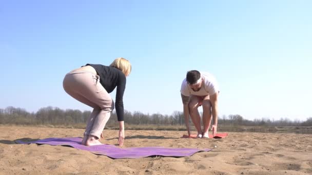 Een blanke vrouwelijke instructeur, een yoga leraar, leert een man met een baard hoe je asana's goed uitvoeren in yoga, een natuur sessie op een rivier en een zandstrand. — Stockvideo