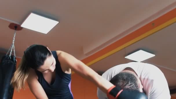A slim girl with fat guy fool and have fun in gym. A woman beats a plump sweat man in boxing gloves. Individual weight loss drills for thick man. Training with personal coach. Workout with instructor — Stock Video