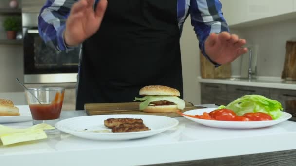 Ο υπέρβαρων άντρας με ποδιά μαγειρεύει χάμπουργκερ στο σπίτι. Όλα τα συστατικά και τα προϊόντα είναι στο τραπέζι. Ανθυγιεινό τρόπο ζωής, τηγανητό και υψηλής θερμιδικής τροφής. Ο κίνδυνος της παχυσαρκίας και του υπέρβαρος. Αργή — Αρχείο Βίντεο