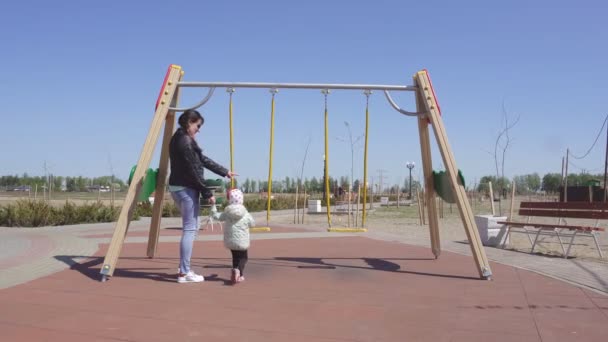 Niedliche kaukasische Mutter rollt ihre kleine Tochter 3 Jahre auf einer neuen Schaukel, Spiel auf dem Spielplatz, bunt — Stockvideo