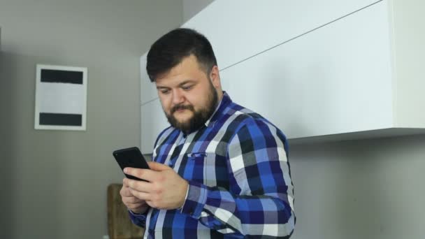 Un hombre regordete con barba mirando el teléfono y volteando. El gordo roba una pantalla del teléfono. Hombre con una taza de café mira noticias en el teléfono inteligente y navega en Internet y charlando con seguidores o suscriptores — Vídeo de stock