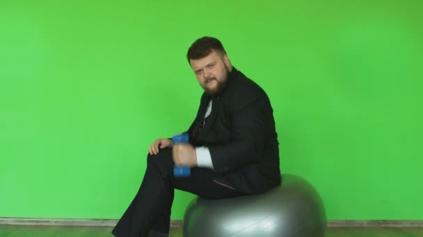 Funny fat affärsman med hantel sitter och rör sig på övning bollen i gymmet. Chromakey grön. Skäggiga tjocka killen i svart jacka, skjorta och slips är i Fitness Club. — Stockvideo