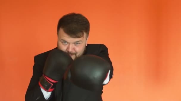 Manager in suit and gloves is boxing and throws punches at the camera in slowmo on orange background. Cheerful plump Chief or positive fat boss relieves stress in sport centre. — Stock Video