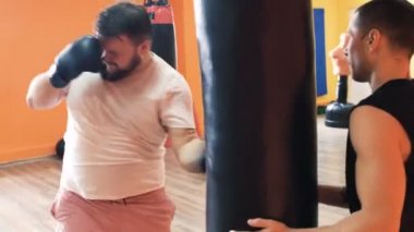 Eldiven içinde şişko adam spor salonunda punç torbası atıyor. Dolgun adam için bireysel kilo kaybı matkaplar. Kişisel antrenörlerle boks eğitimi. Bir spor salonunda eğitmen ile egzersiz. Kalın için özel eğitmen