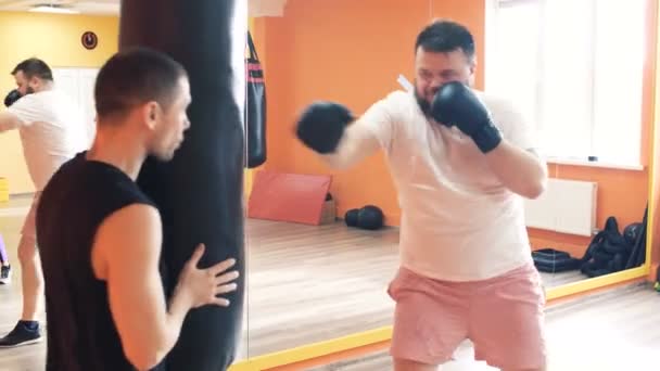 Thick man in gloves beats punching bag in gym. Individual weight loss drills for plump guy. The boxing training with personal coach. Workout with instructor in a sport hall. Private trainer for thick — Stock Video