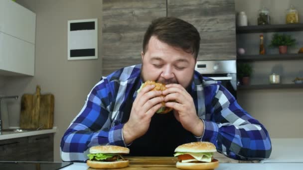 Dikke kerel eet en geniet van zijn hamburger. Persoon met een baard eet voedsel dat hij kookte. Ongezonde levensstijl, gebakken en hoge calorie schadelijk voedsel. Het risico van obesitas en overgewicht. Slow Motion. — Stockvideo