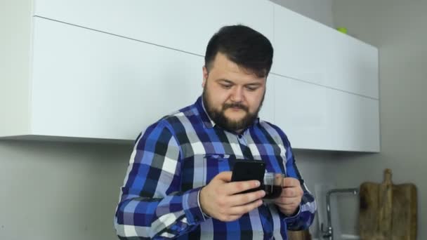 Толстяк стоит на кухне, смотрит на телефон и пьет кофе. Большой мужчина на кухне смотрит и давит на смартфон и пьет кофе. . — стоковое видео