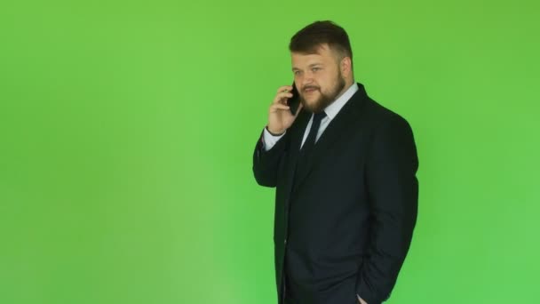 Бородатый толстый бизнесмен в черном костюме говорит по телефону. Зеленый хромакей. Толстяк решает проблемы с помощью смартфона. Человек обсуждает важные вопросы — стоковое видео