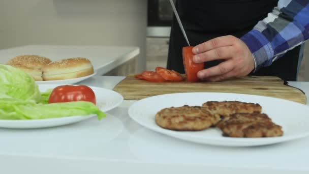 Chico regordete cocina hamburguesas caseras en su cocina. El hombre corta tomates en la tabla de cortar. Estilo de vida poco saludable, alimentos fritos y nocivos con alto contenido calórico. El riesgo de obesidad y sobrepeso. Movimiento lento . — Vídeo de stock